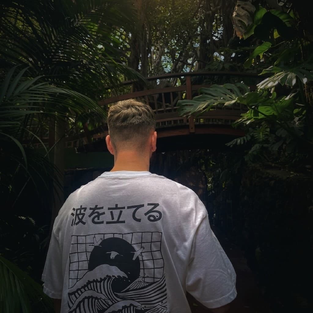 Tyler Hammond photographed from behind in softly dim, canopy-filtered light among deep green tropical plants, wearing an oversized white T-shirt printed with the Japanese characters 波を立てる (meaning “make waves”) and a black graphic of waves, flying cranes, and a large circle in the style of Japanese woodblock art; a wooden footbridge is visible through the trees behind him.