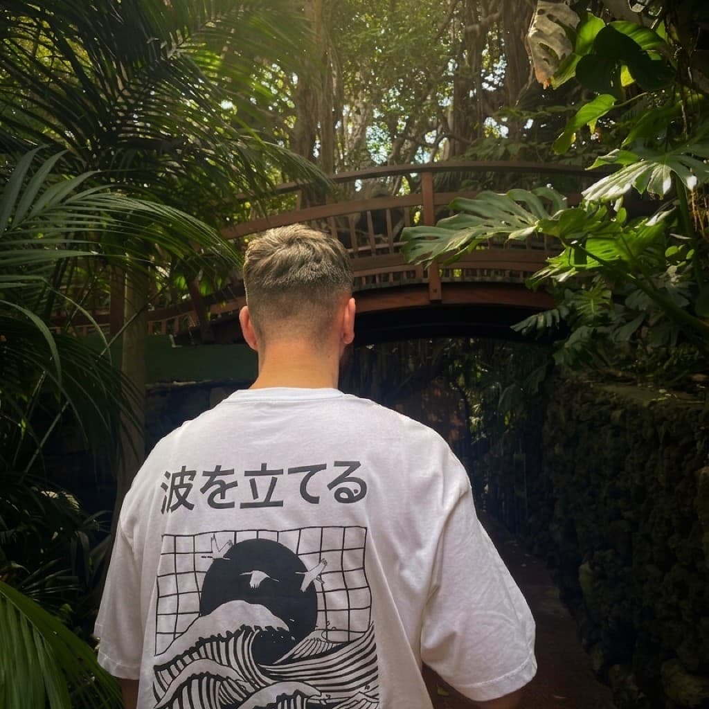 Tyler Hammond photographed from behind in bright, sunlit tropical greenery, wearing an oversized white T-shirt printed with the Japanese characters 波を立てる (meaning “make waves”) and a black graphic of waves, flying cranes, and a large circle in the style of Japanese woodblock art; a wooden footbridge is visible through the leaves ahead.