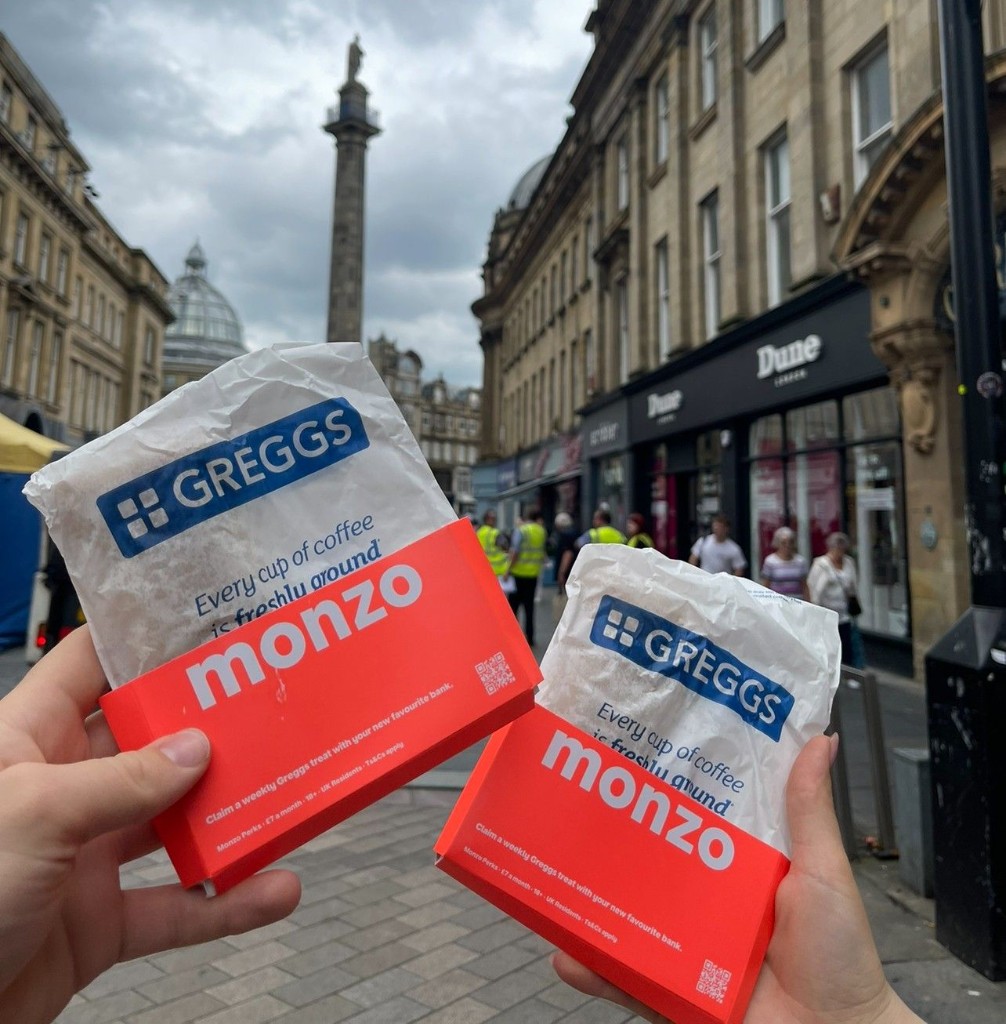 Two hands holding Greggs bags in coral Monzo Perks sleeves on Grey Street in Newcastle, with Grey’s Monument visible in the background.