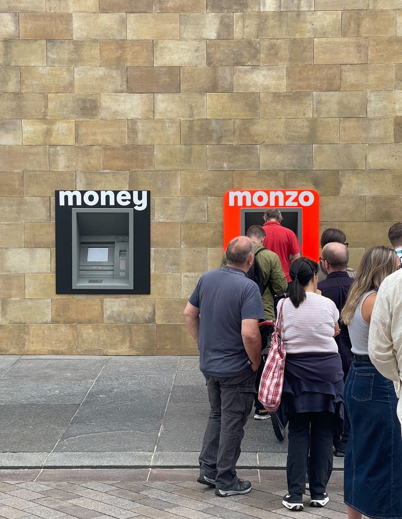 Side-by-side ATMs on Grainger Street labelled “money” and “monzo”: a queue waits at the coral Monzo sausage-roll ATM while the generic money ATM stands unused.